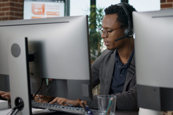 African american employee using headset at customer service job, asnwering call about telemarketing sales. Male operator working at call center office to help clients on helpline.